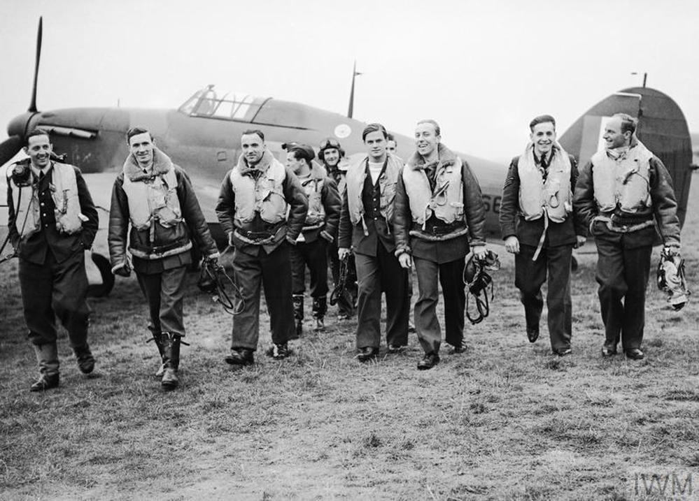 Polish WW2 Battle of Britain pilots of 303 Squadron in flying gear walk towards the camera in front of parked Hawker Hurricane aircraft.