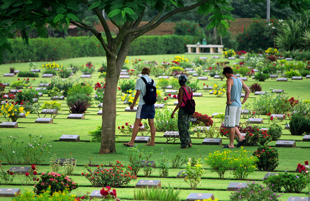 Visitors to Chungkai War Cemetery observe bronze headstone plaques set amongst a vivid green lawn dappled with yellow and red flowering plants in headstone borders.