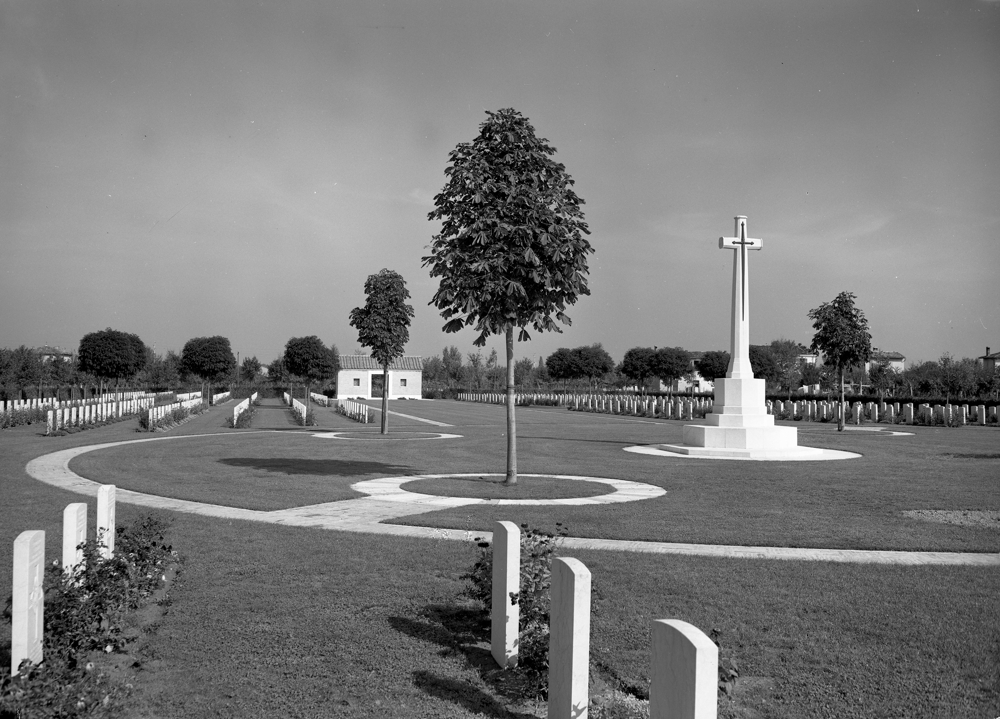 Faenza War Cemetery with central Cross of Sacrifice set among a circular path around which rows of CWGC headstones have been planted. A Pair of trees is visible in the foreground while the cemetery shelter building is visible at the back of the cemetery.