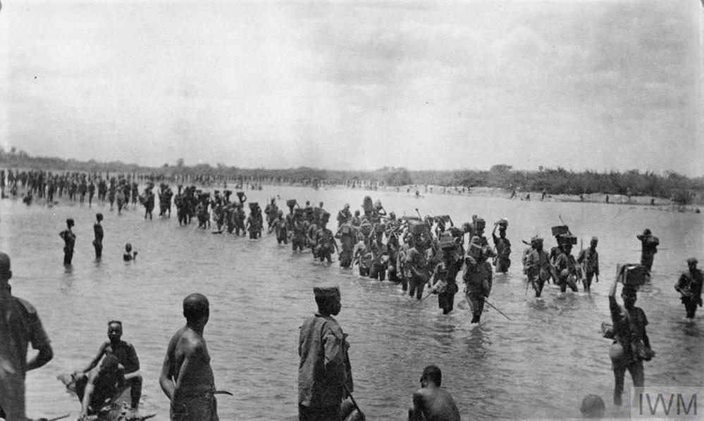 Soldiers, porters, and carriers of the King's African Rifles wading through a low river.